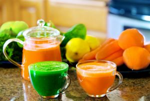Two cups of fresh vegetable juice on kitchen counter with vegetables in background