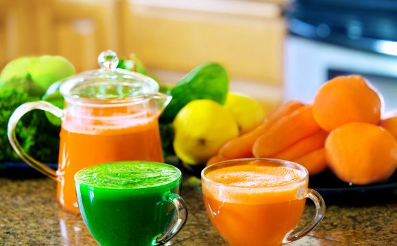 Two cups of fresh vegetable juice on kitchen counter with vegetables in background