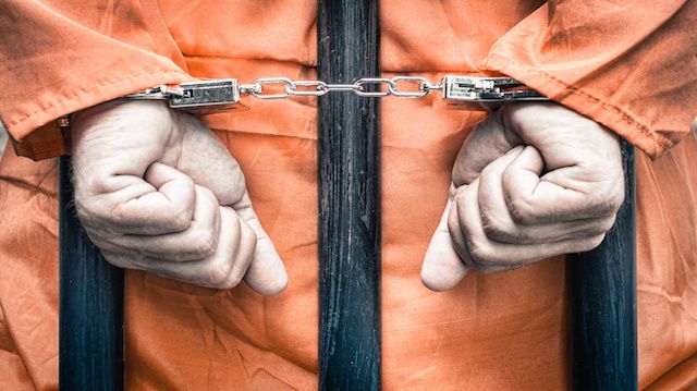 Handcuffed hands of a prisoner behind the bars of a prison with orange clothes - Crispy desaturated dramatic filtered look