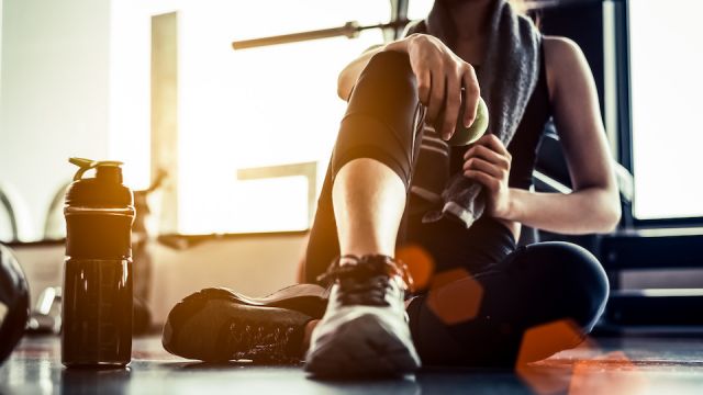 Sport woman sitting and resting after workout or exercise in fitness gym with protein shake or drinking water on floor. Relax concept. Strength training and Body build up theme. Warm and cool tone