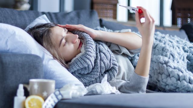 Sick exhausted girl in scarf is lying in bed wrapped in blanket. Young woman with fever and headache is measuring temperature with thermometer, treated at home. Winter cold and flu concept.