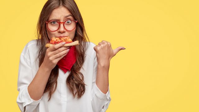 Horizontal shot of surprised female eats tasty slice of pizza, dressed in fashionable clothes, indicates with thumb, invites you to pizzeria, isolated over yellow studio wall. People and nutrition