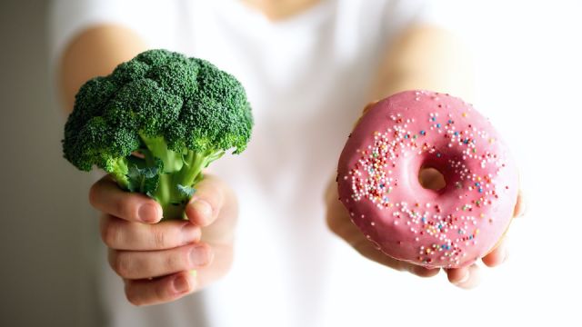 Young woman in white T-shirt choosing between broccoli or junk food, donut. Healthy clean detox eating concept. Vegetarian, vegan, raw concept. Copy space